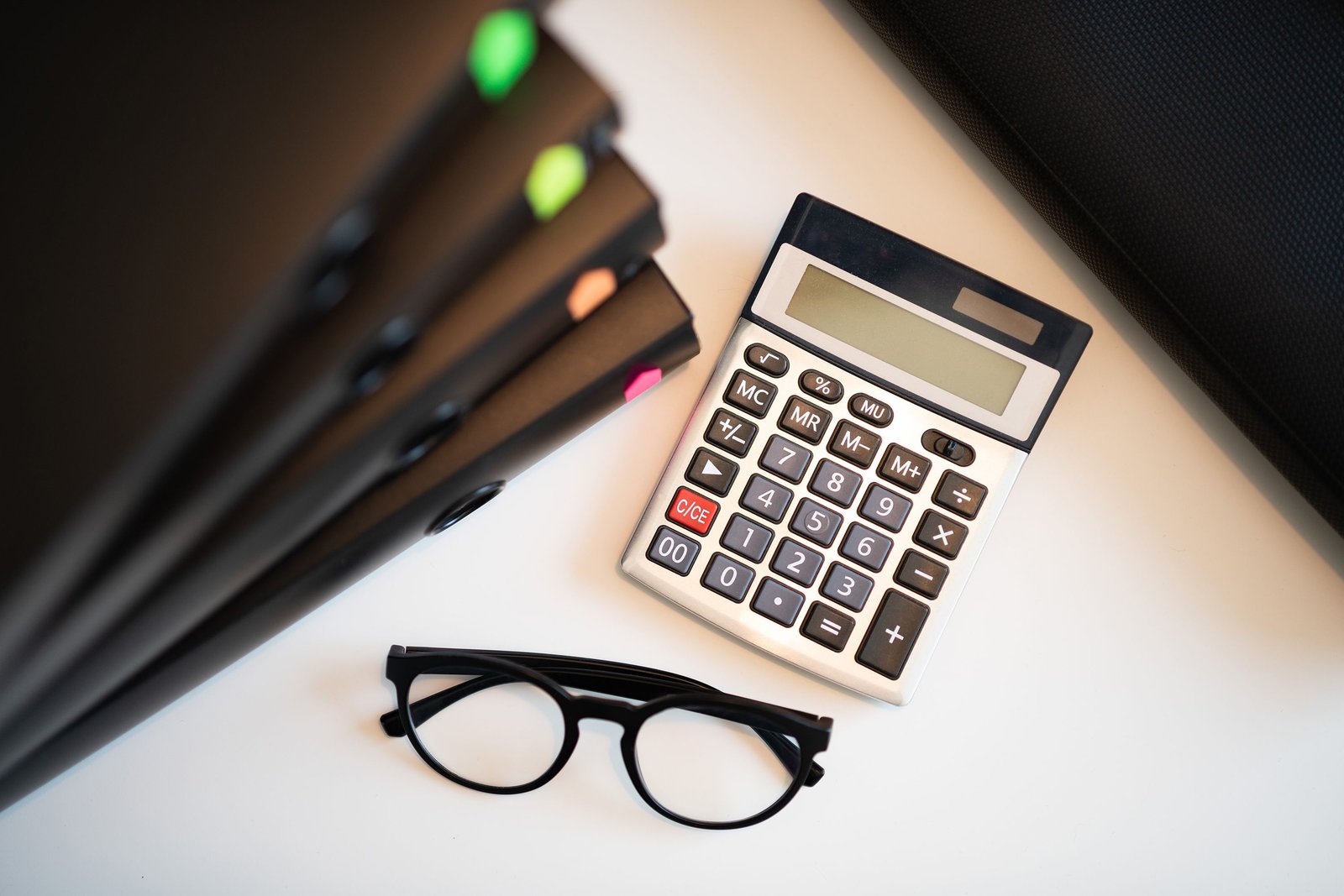 Calculator, eyeglasses, and black folders on a white desk.