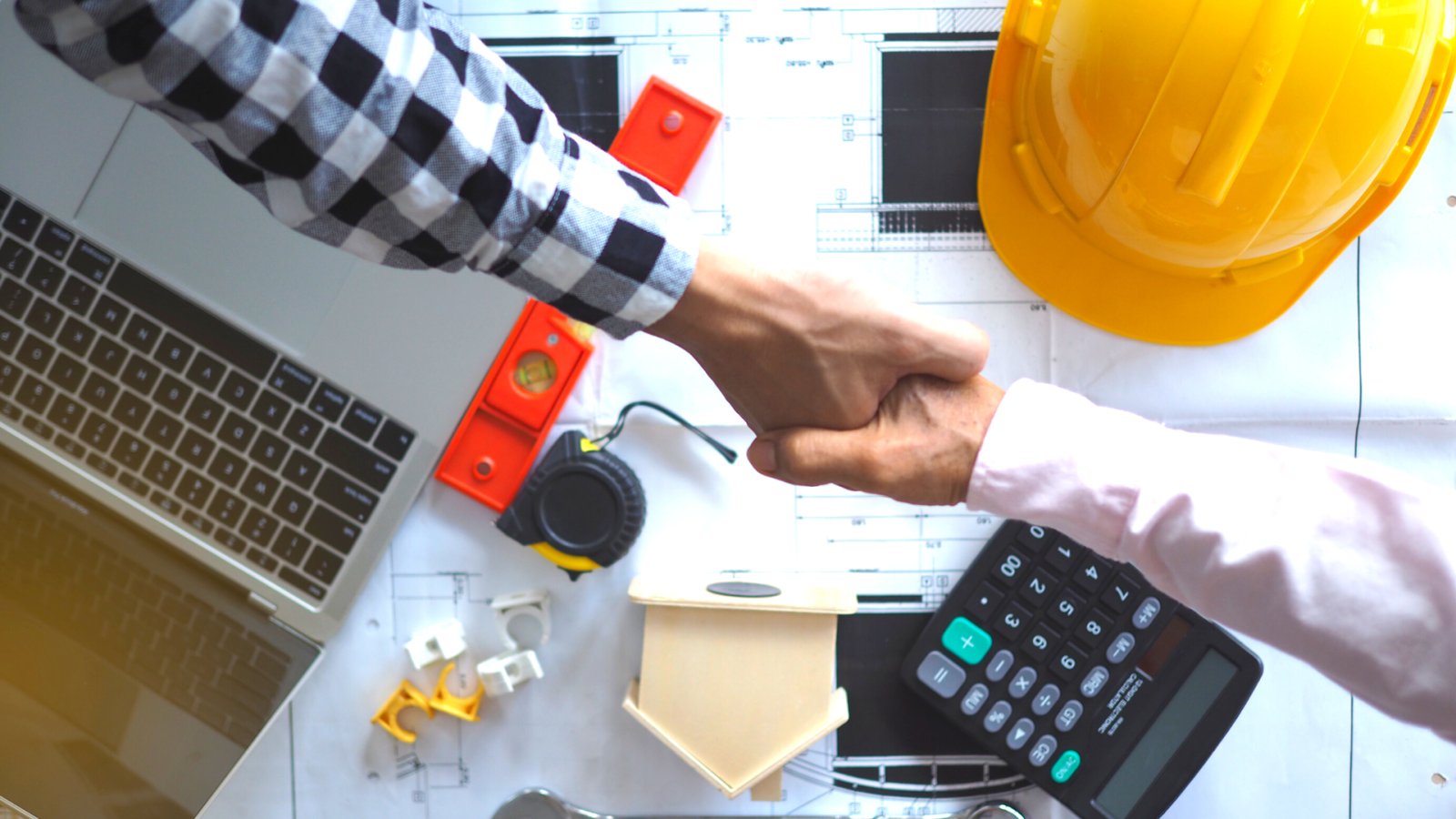Two people shaking hands over construction plans beside a hard hat, laptop, and calculator.