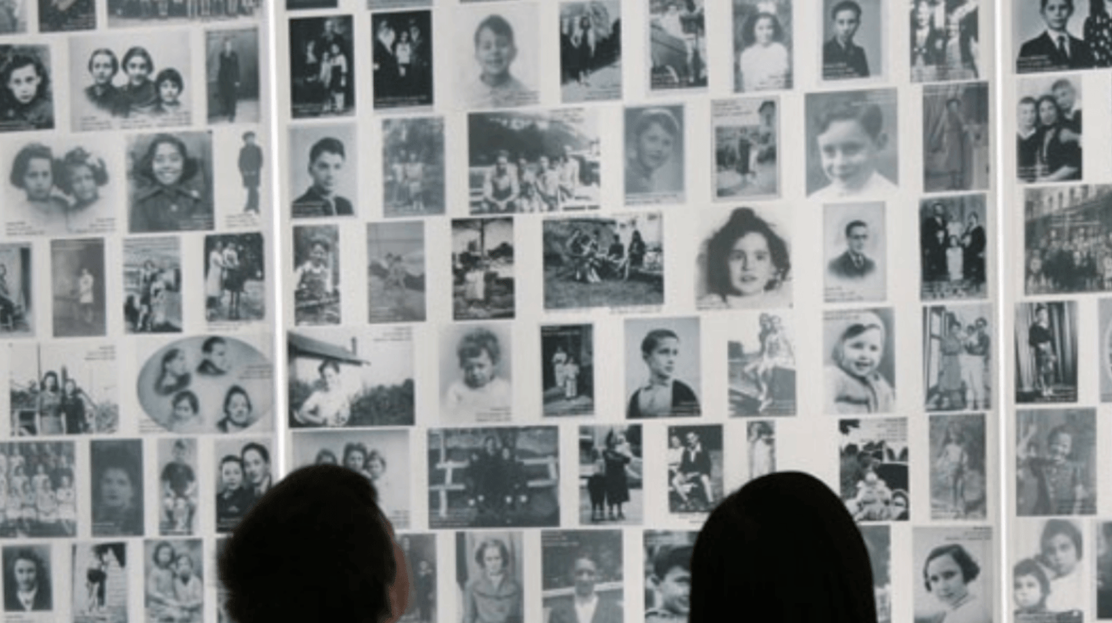 A wall of black-and-white portrait photographs, with two visitors seen from behind.