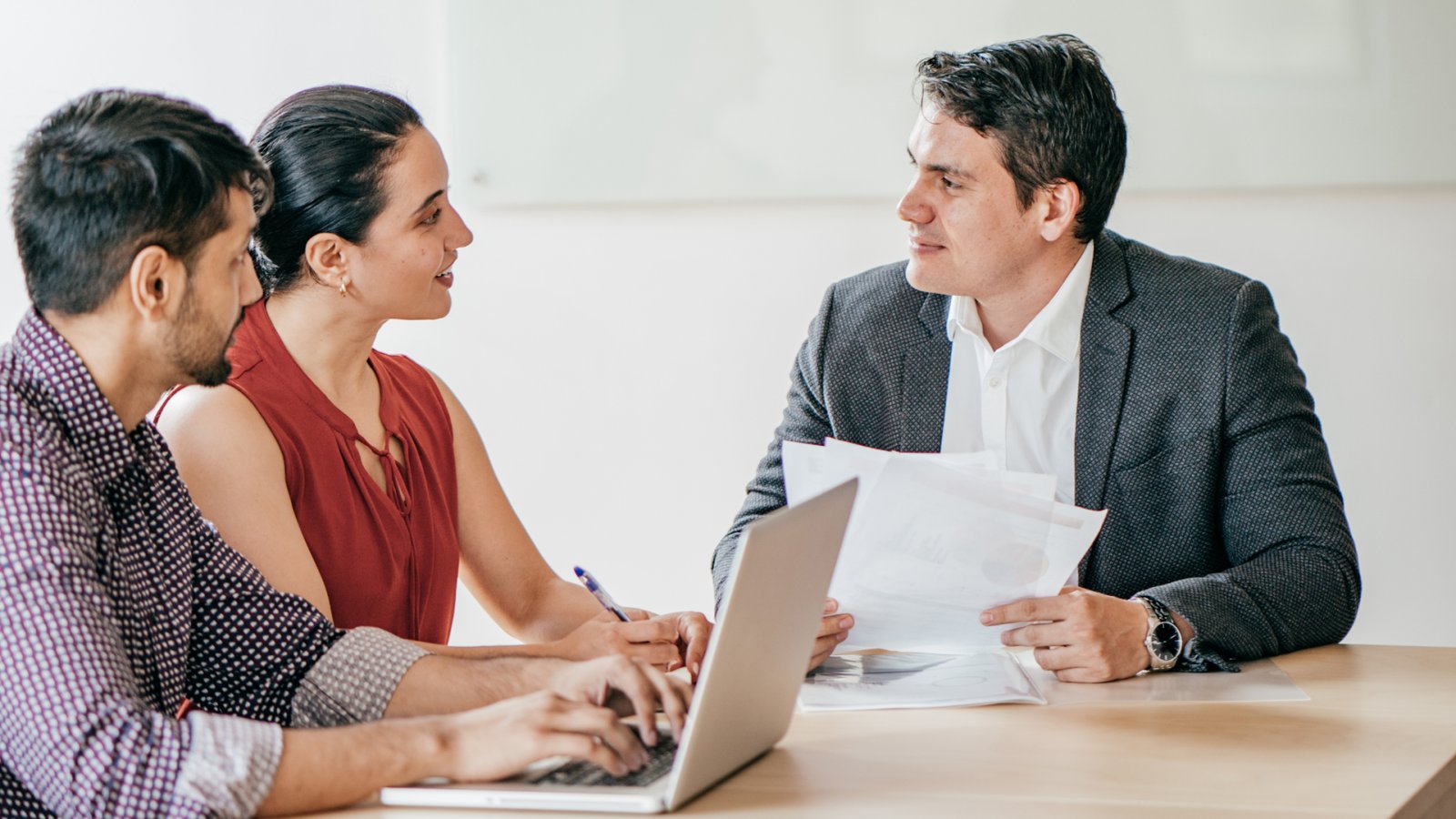 A man holding documents talks with a woman holding a pen and a man holding a tablet at a table.