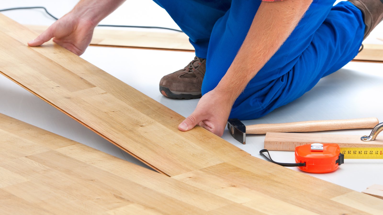 A person fitting a wood-look flooring plank beside a hammer and tape measure.