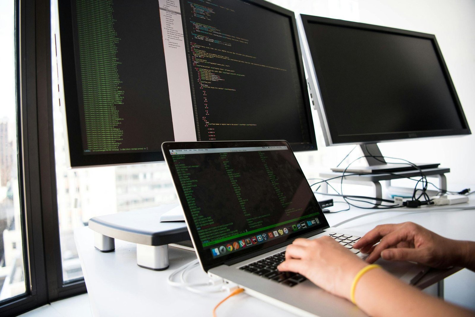 Hands at a laptop with code displayed on the laptop and desk monitors.