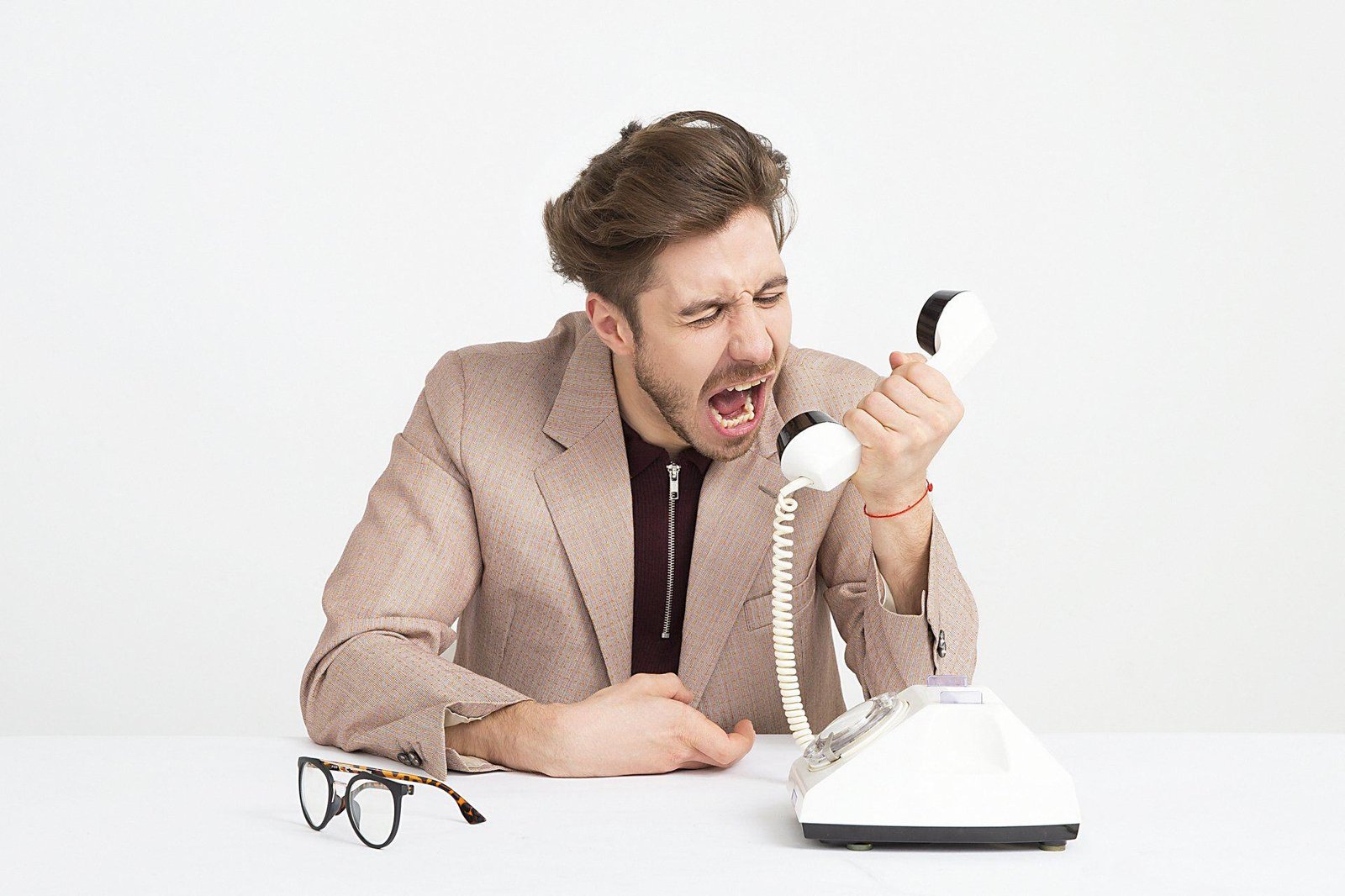 A person holds a telephone handset at a desk beside papers and eyeglasses.