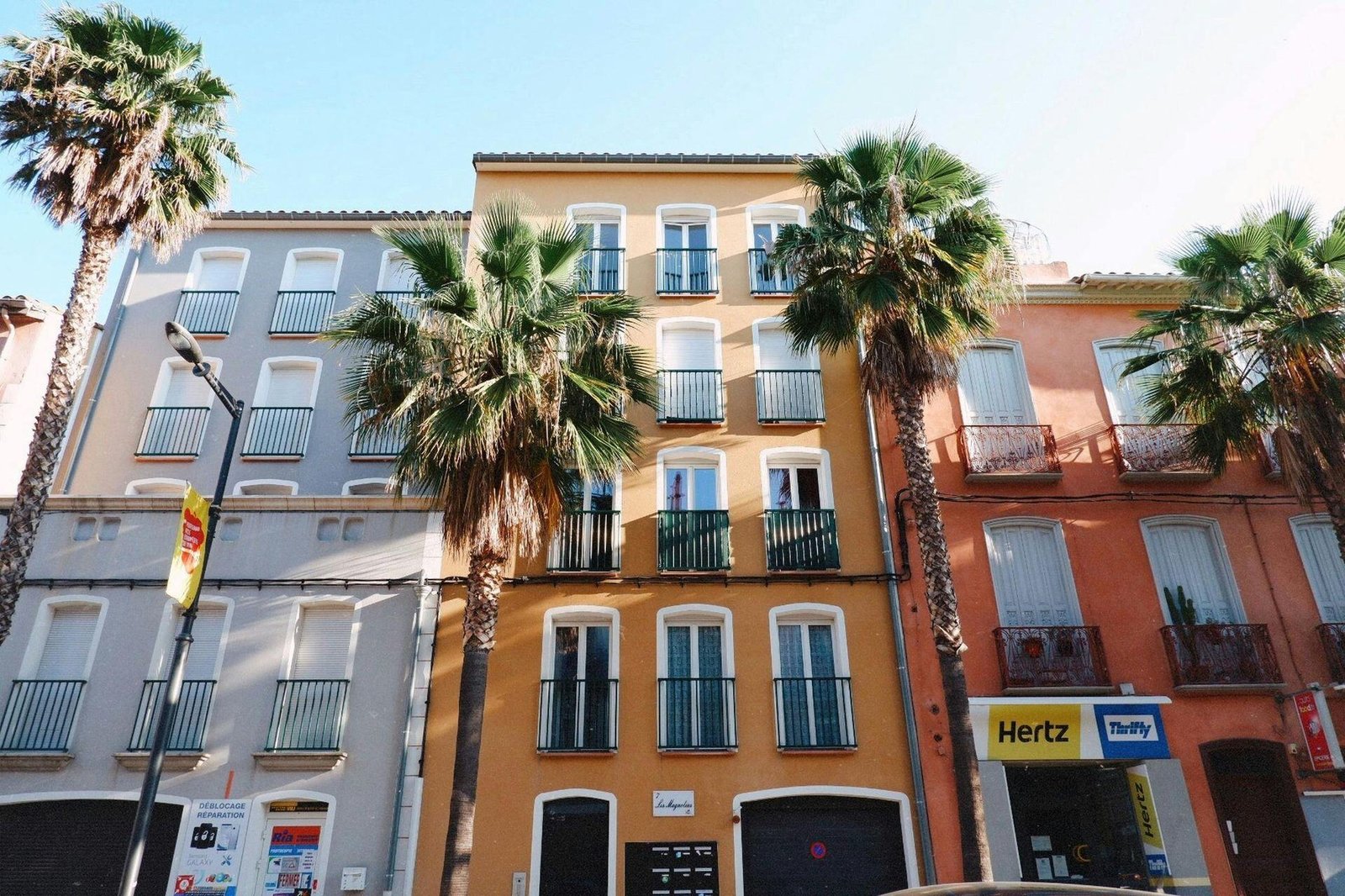 Palm trees in front of multistory buildings with balconies and pale blue, yellow and pink facades.