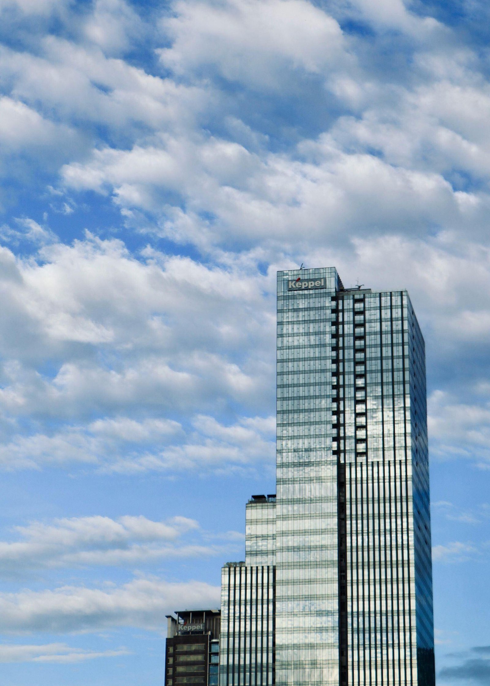 Glass office tower with a rooftop Keppel sign against a cloudy sky.