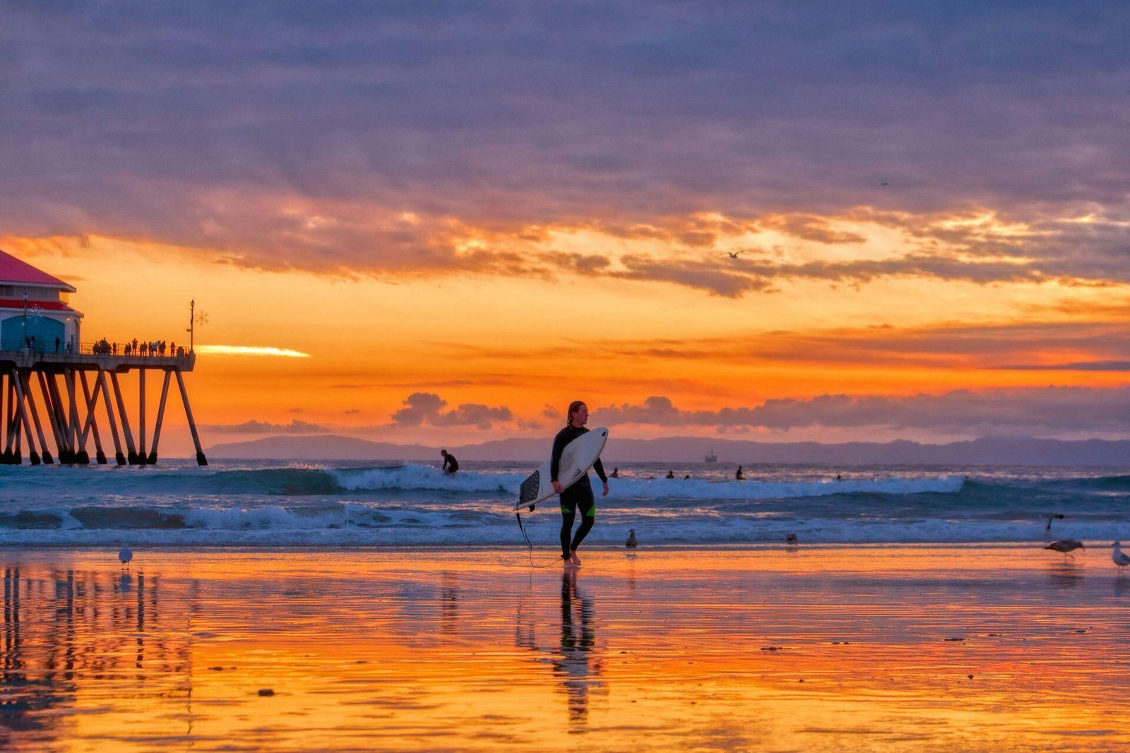 Surfer carrying a surfboard on a beach beside a pier at sunset.
