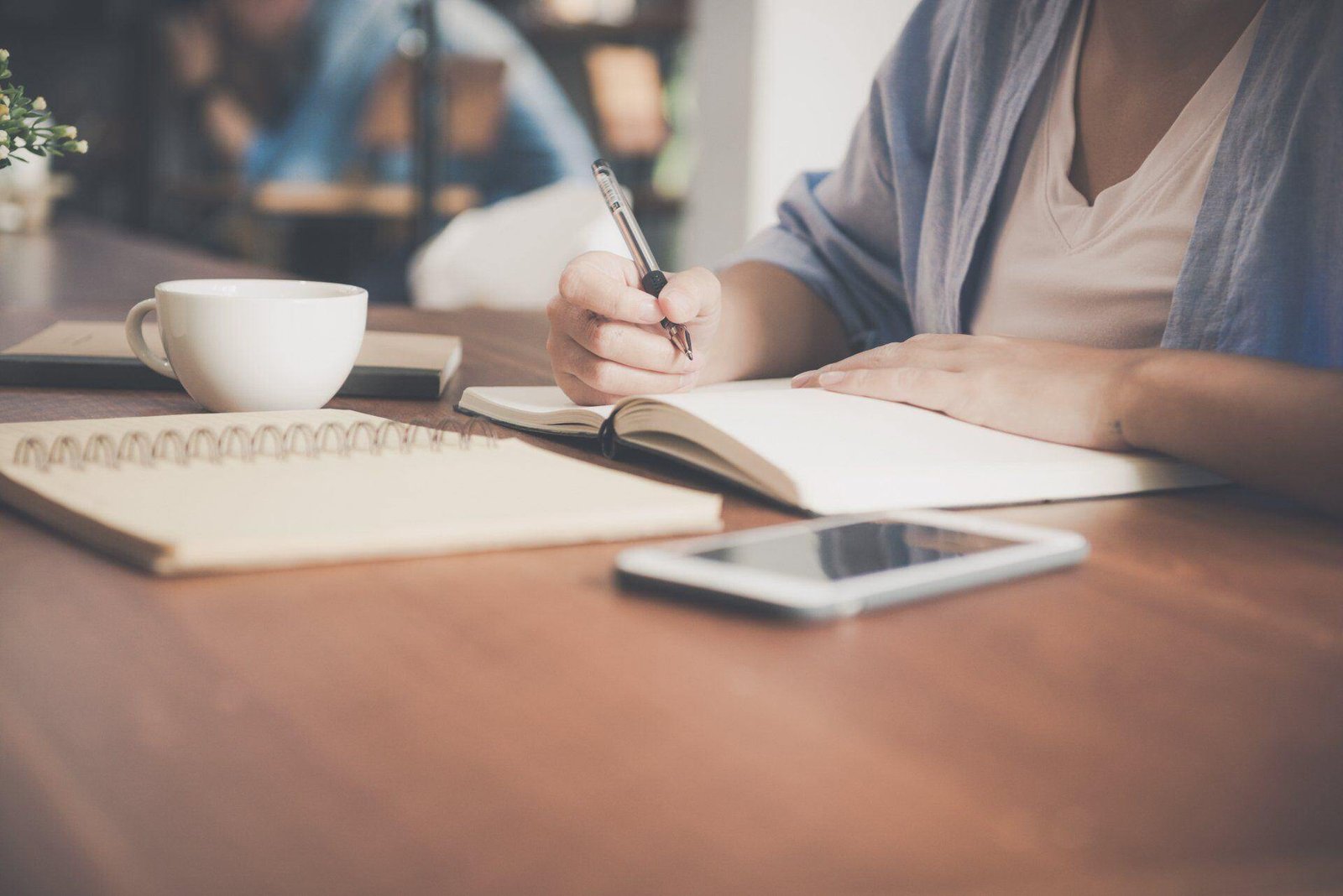 A person writing in a notebook at a table with a mug and smartphone.