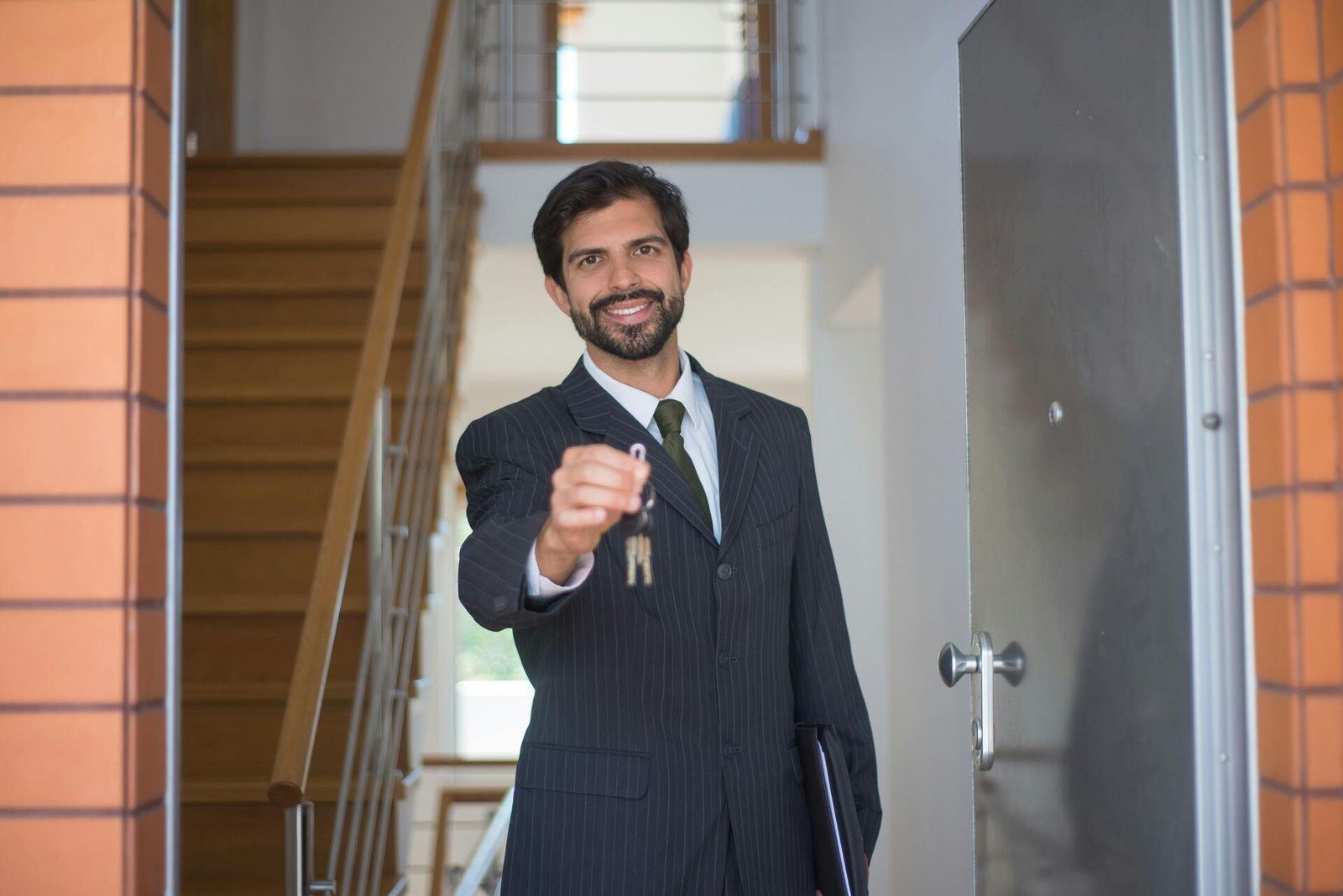 A man in a dark suit holds out a set of keys at an open doorway.