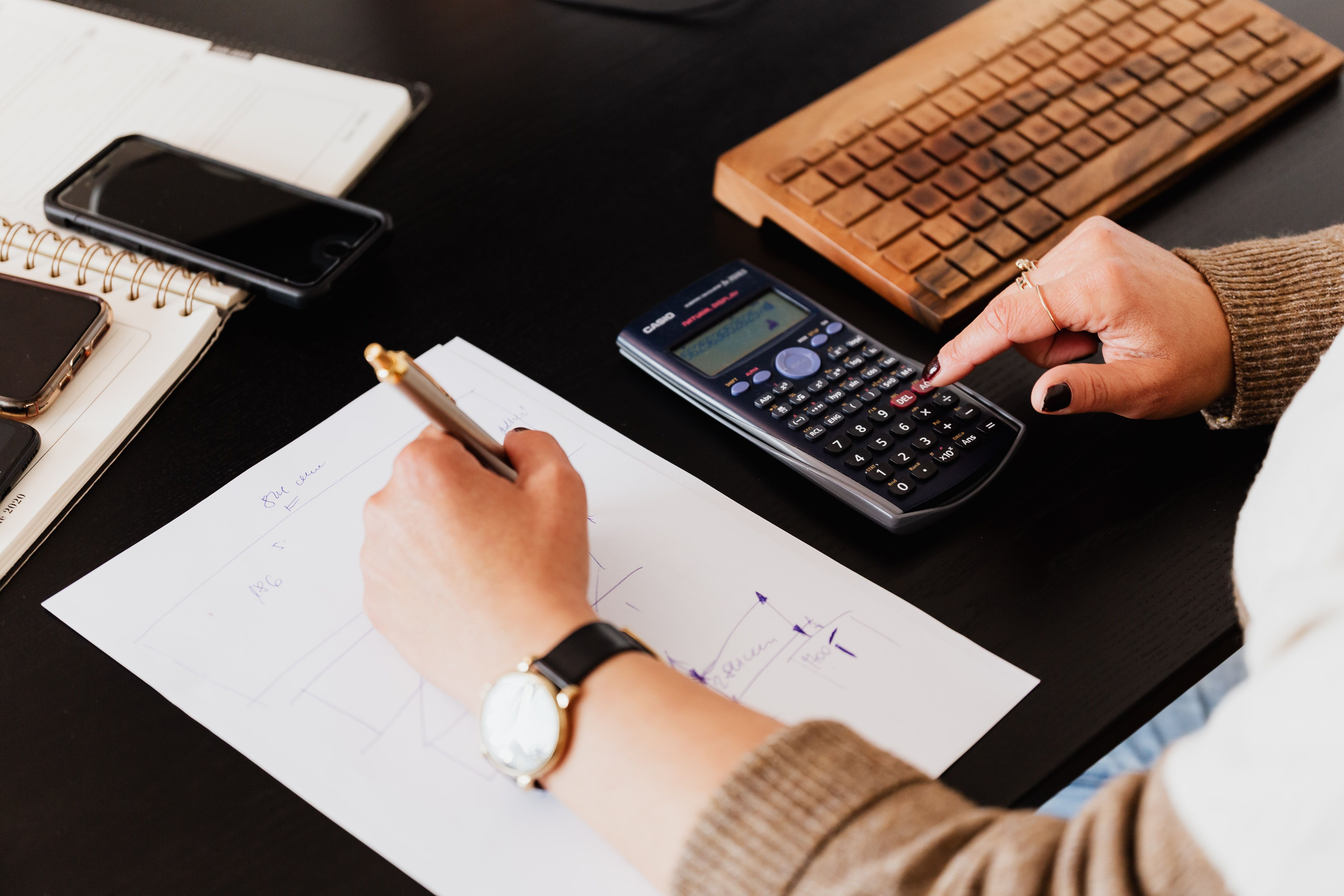 Person writing figures on paper while using a calculator at a desk.