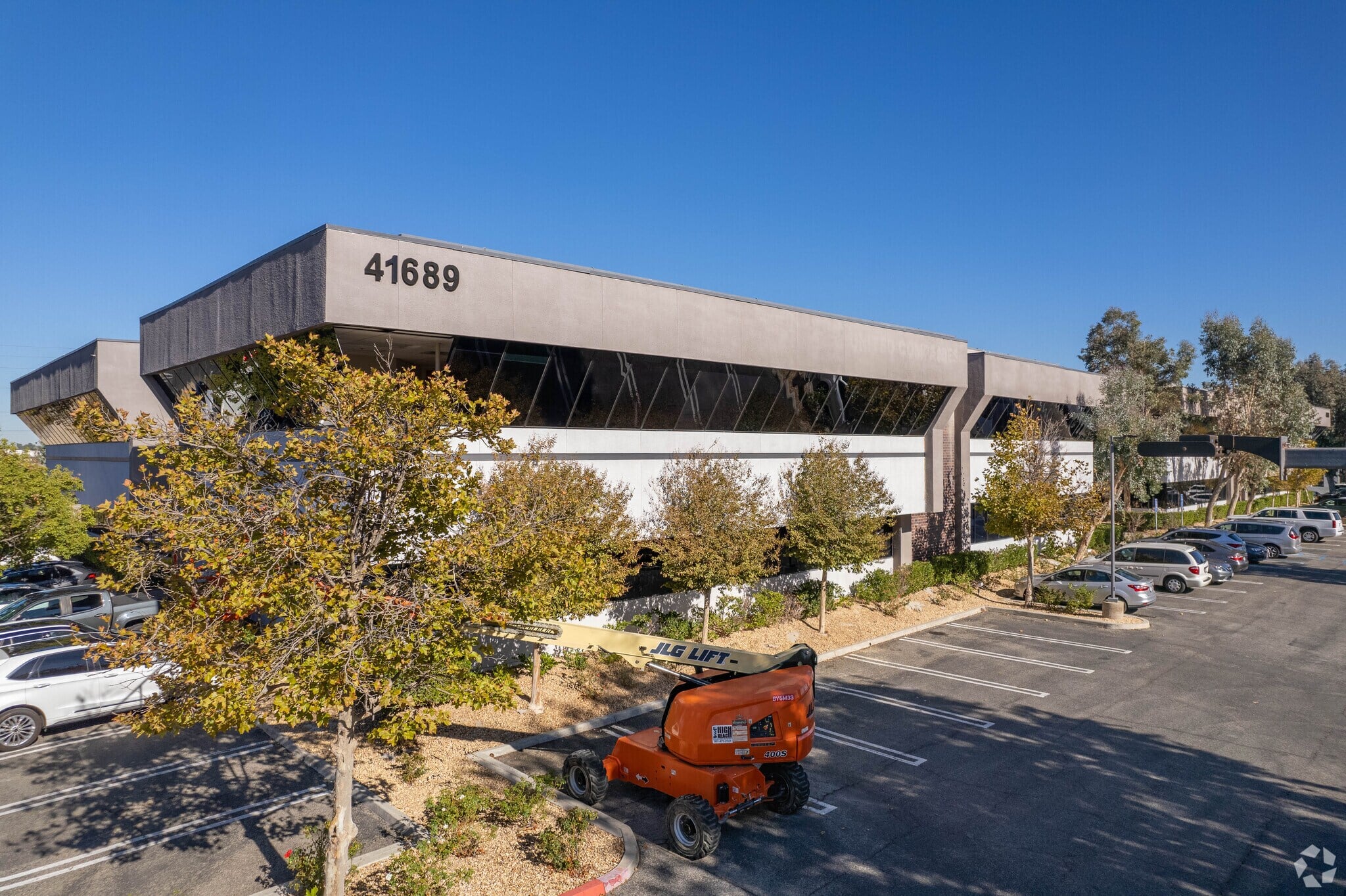 Low-rise office building marked 41689 with mirrored windows, trees, parked cars, and an orange boom lift in the parking lot under a clear blue sky.