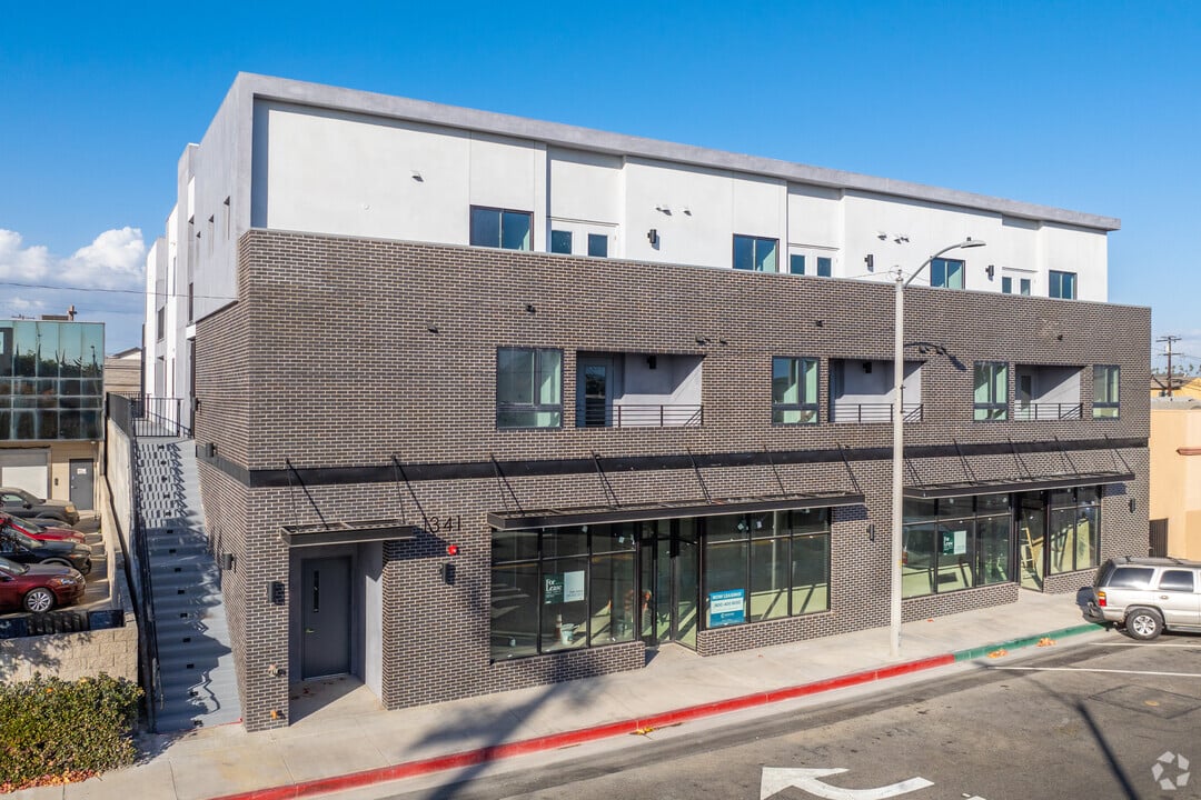Exterior of a three-story mixed-use building with dark brick lower floors, a white upper facade, storefront windows, and street parking under a clear blue sky.