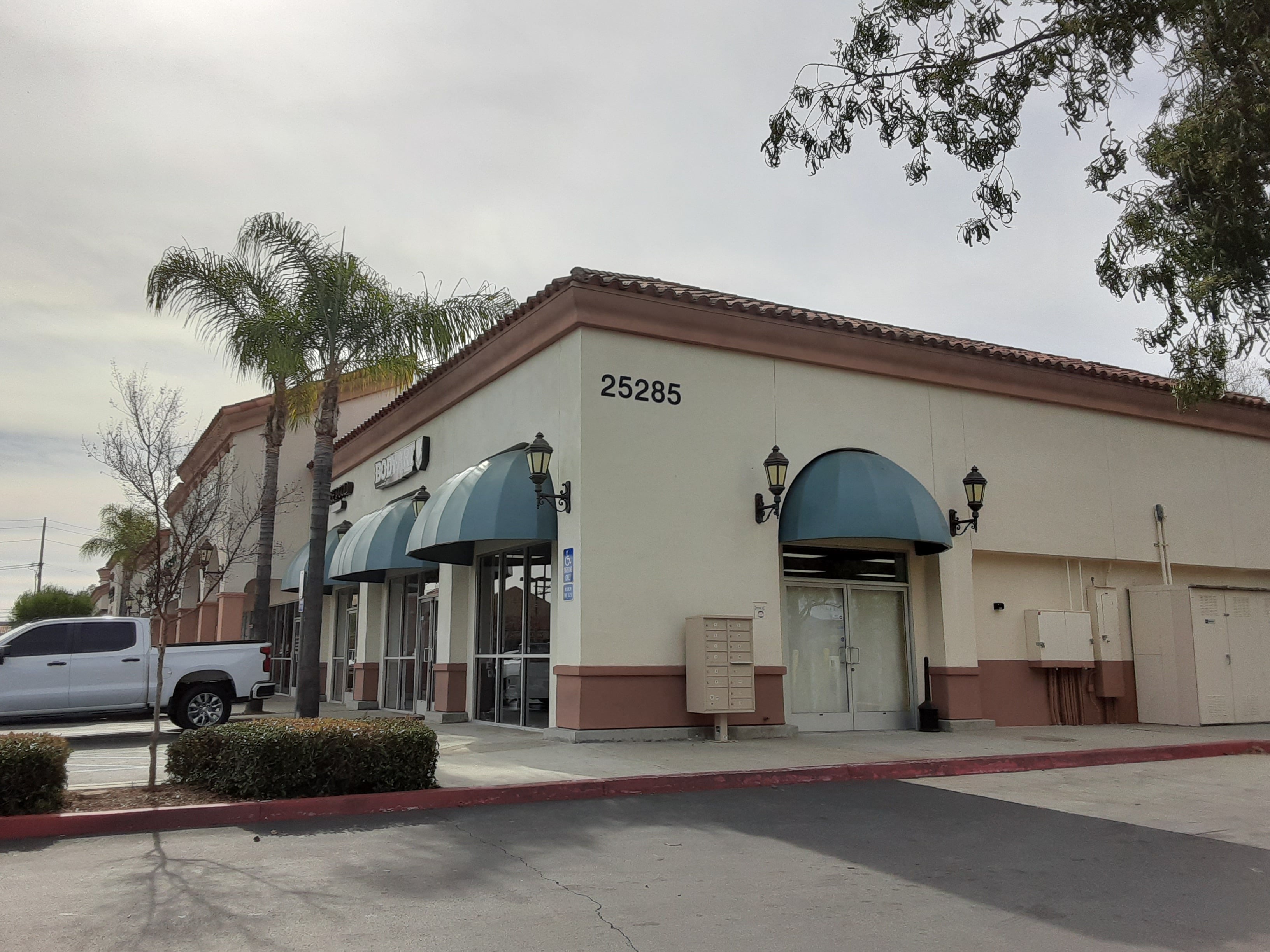 Commercial building exterior with a tile roof, storefront awnings, exterior lighting, utility cabinets, and a paved drive under an overcast sky.